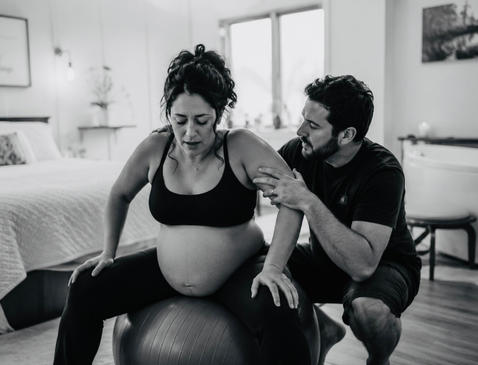 Partner supporting a laboring mother on a birth ball in a calm, candle-lit birth room.